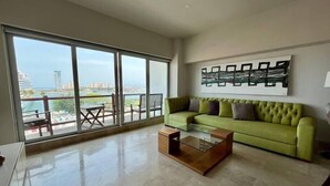 Ein geräumiges Wohnzimmer mit einem grünen Sofa, einem Glastisch und einem großen Fenster mit Blick auf eine Stadtlandschaft und ein Gewässer.