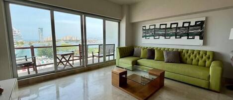 Ein geräumiges Wohnzimmer mit einem grünen Sofa, einem Glastisch und einem großen Fenster mit Blick auf eine Stadtlandschaft und ein Gewässer.