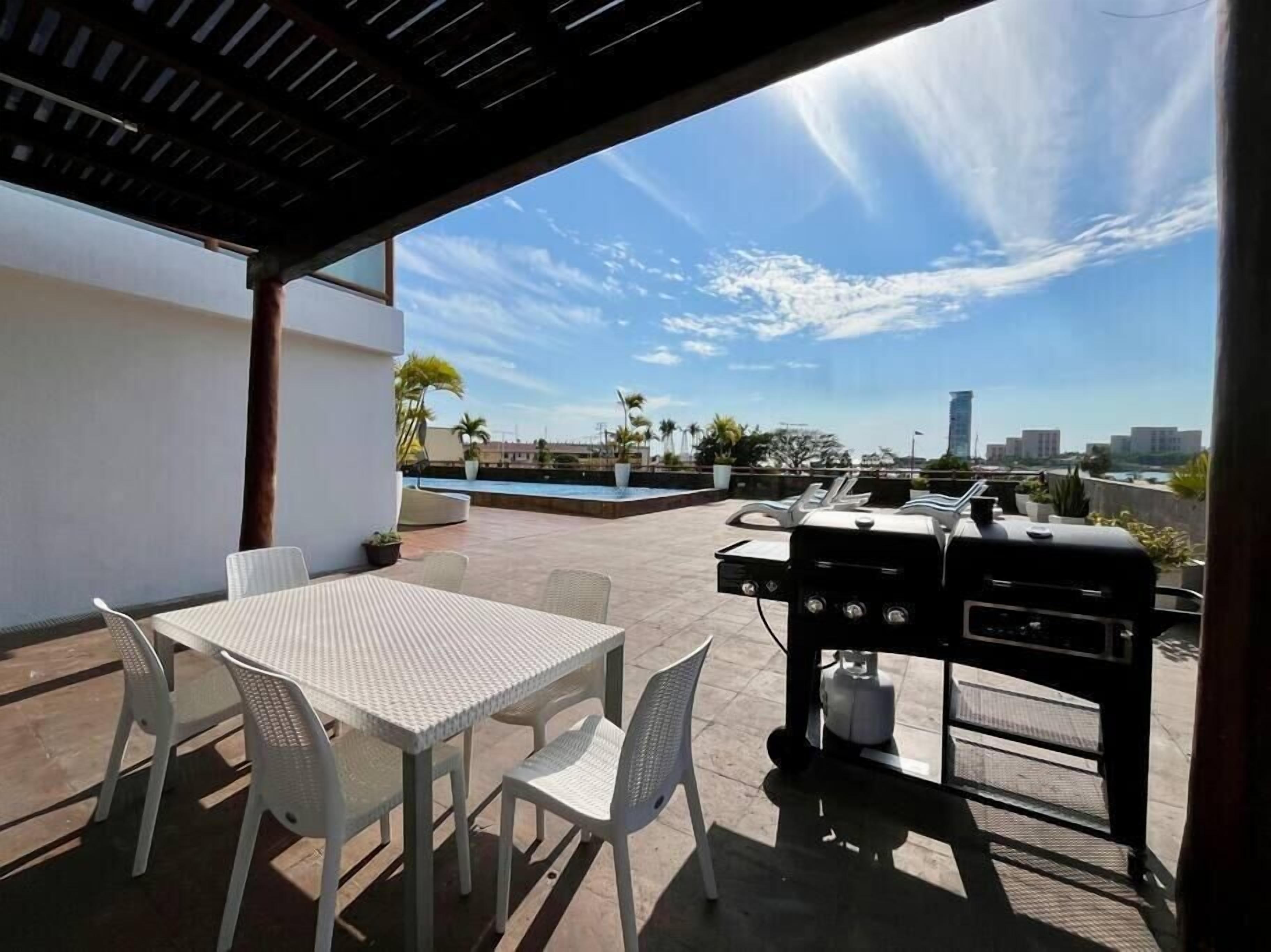 An outdoor patio with a table and chairs, a grill, and a swimming pool in the background.