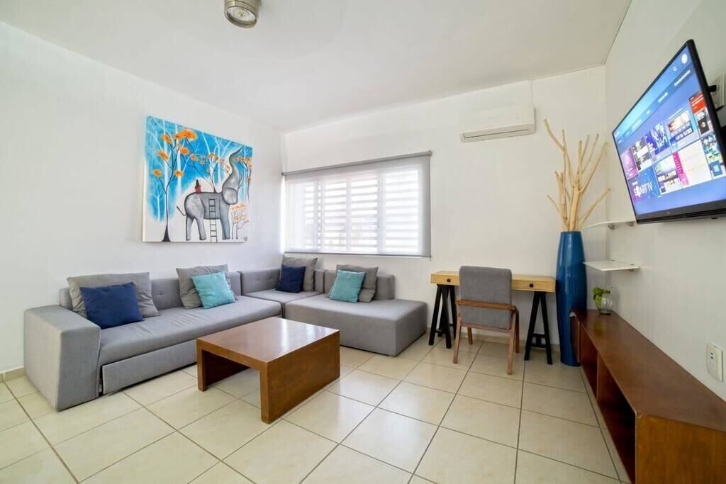 A modern living room with a grey sectional sofa, a wooden coffee table, a desk with a chair, and a flat-screen TV mounted on the wall.