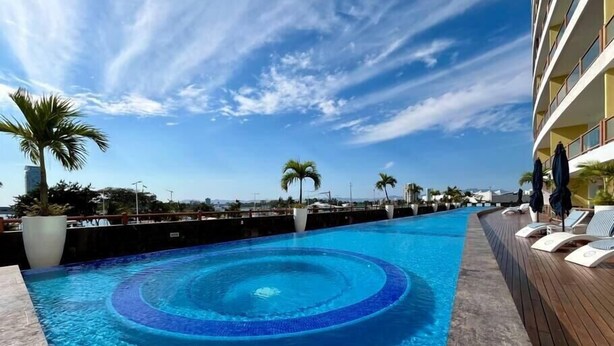 Ein Pool mit kreisförmigem Design, umgeben von Liegestühlen und Palmen, mit blauem Himmel und Gebäuden im Hintergrund.