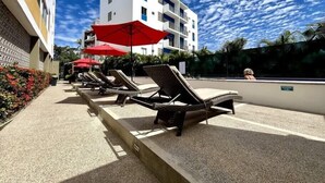 A poolside area with lounge chairs and red umbrellas, a white building in the background, and a clear sky.