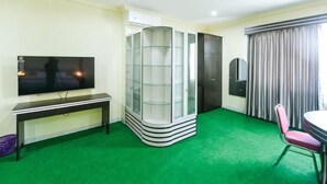 A hotel room with a flat-screen TV, a glass-fronted storage unit, a green carpet, a dining table with a chair, and a window with curtains. - RedDoorz @ Brother Homestay Kupang (Kupang)