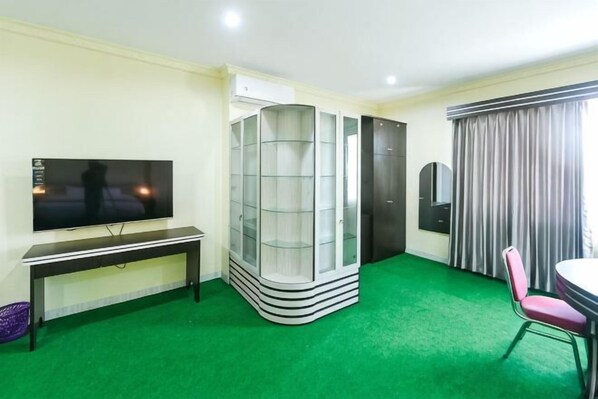 A hotel room with a flat-screen TV, a glass-fronted storage unit, a green carpet, a dining table with a chair, and a window with curtains. - RedDoorz @ Brother Homestay Kupang (Kupang)