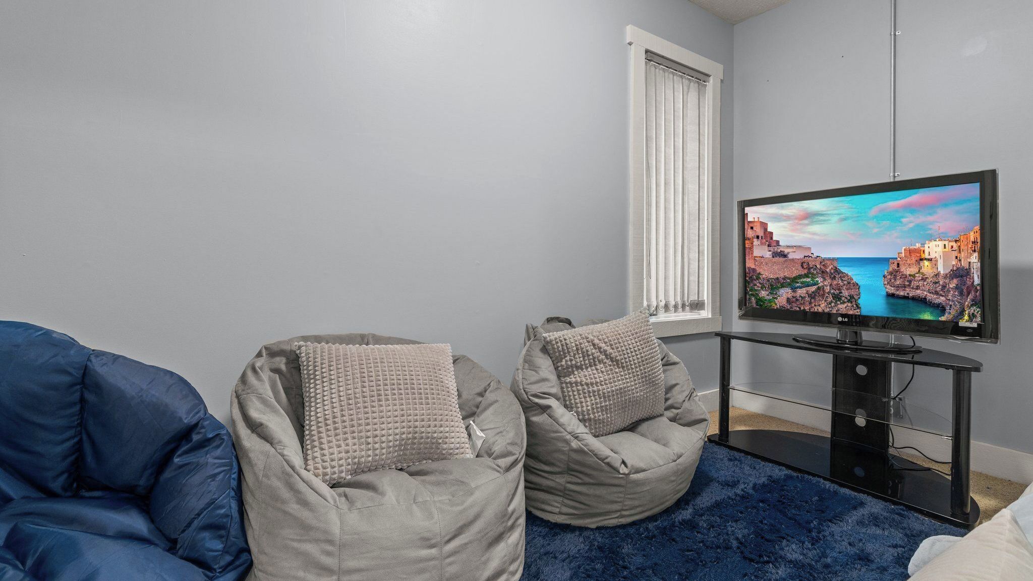 Ein Wohnzimmer mit Fernseher, zwei Sitzsäcken, einem blauen Teppich und einem Fenster mit Jalousien.