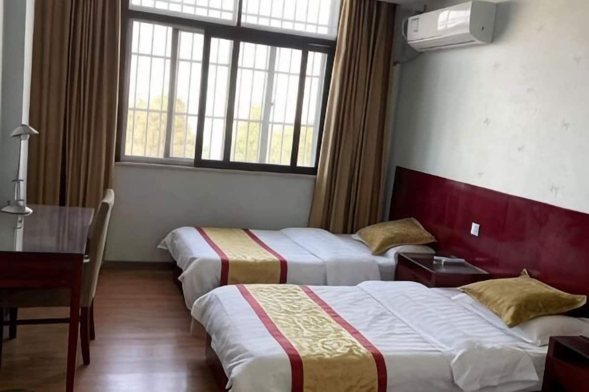 A hotel room with two beds, a desk, a chair, a window with curtains, and an air conditioning unit.