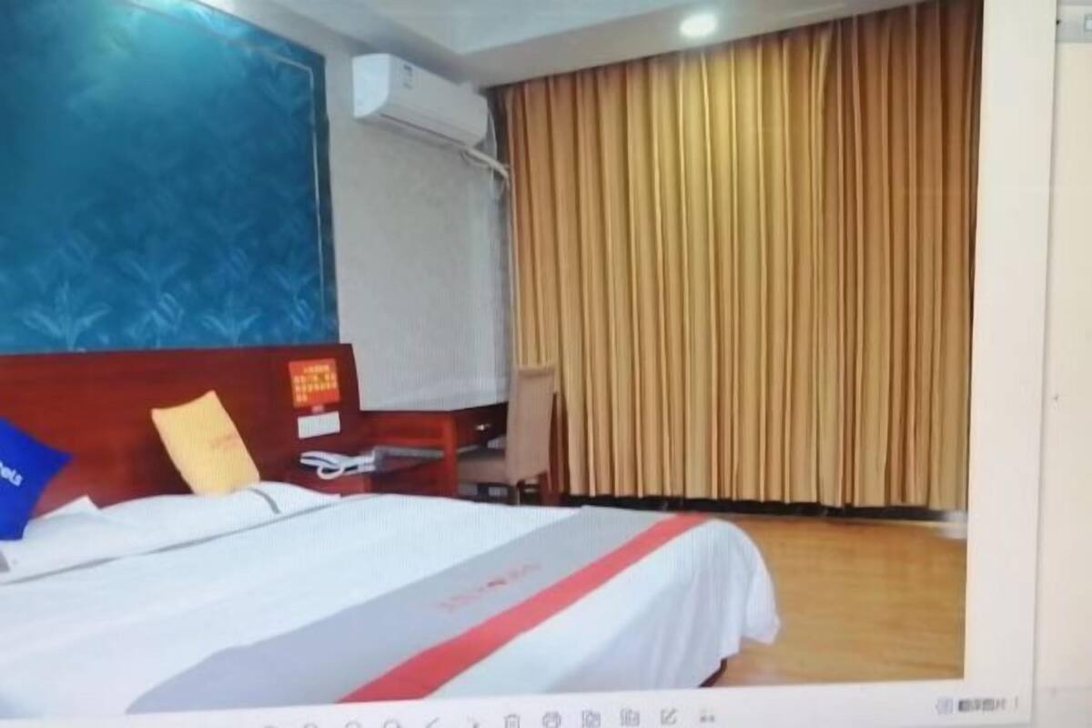 A hotel room with a bed, a desk, a chair, a window with curtains, and an air conditioning unit.