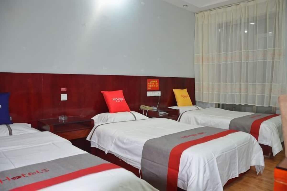 A hotel room with three beds, each with a red and white bedspread, a wooden headboard, and a large window with sheer curtains.