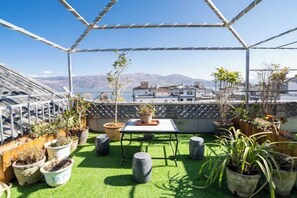 Een dakterras met een geometrische metalen constructie, plantenbakken, een klein tafeltje en uitzicht op de stad en bergen.