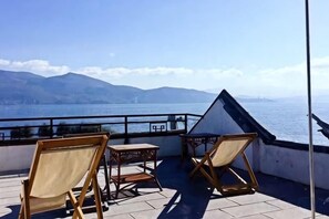 A terrace with two wooden chairs and a small table, overlooking a body of water and distant mountains.