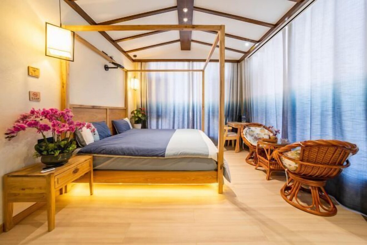 Ein Schlafzimmer mit einem hölzernen Himmelbett, einem Nachttisch mit Lampe, einem Sessel und einem kleinen Tisch mit Pflanze.