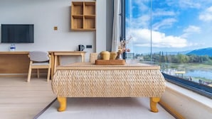 A modern room with a large window, a wooden desk, a television, and a woven ottoman.