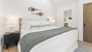 A neatly arranged bedroom with a large bed, bedside lamps, a wooden nightstand, and a door. - Creekside 3B/3B Townhome - 1 Block to Ski Lift! (Park City)
