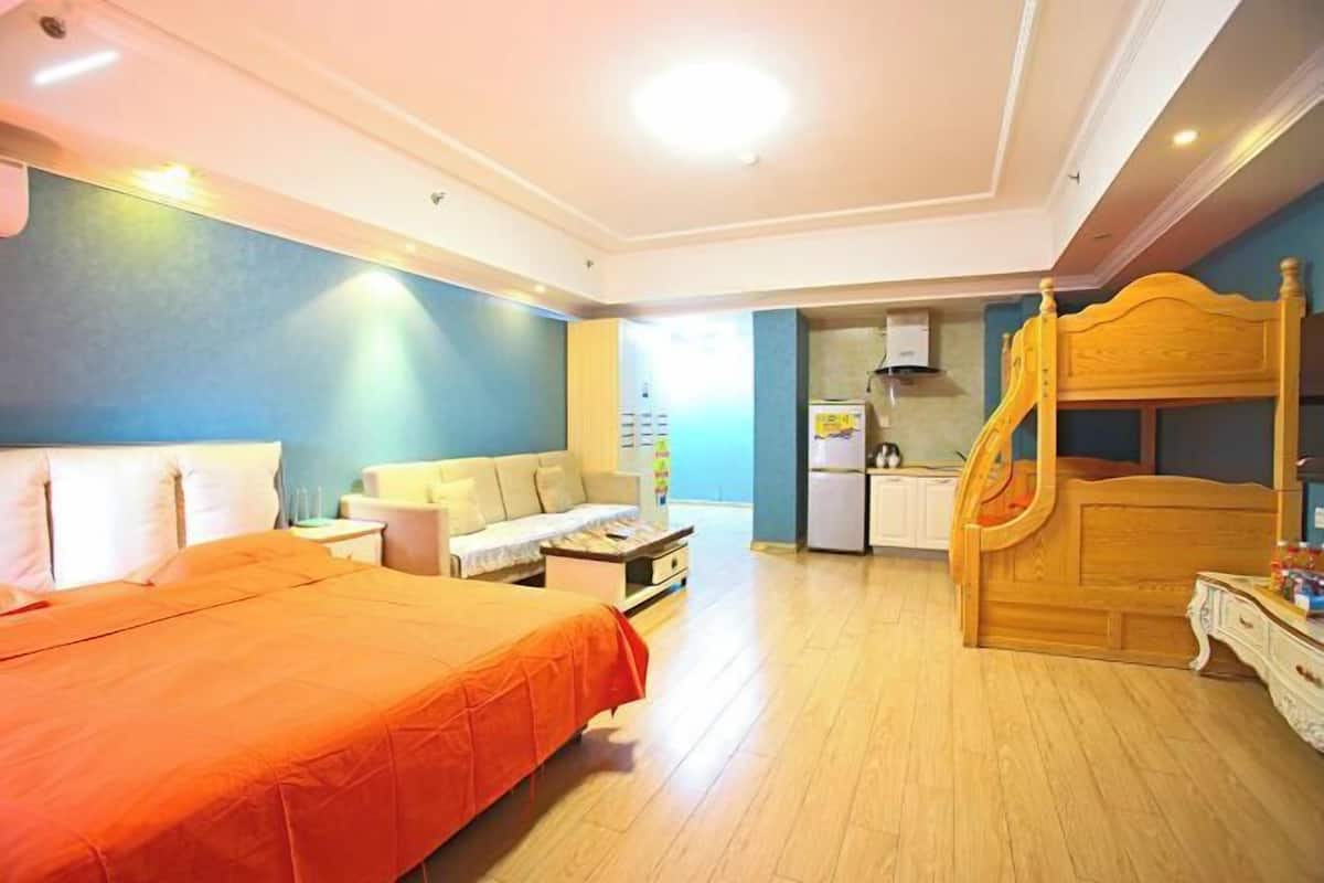 A bedroom with a bed, a wooden headboard, a small desk, and a kitchenette area.