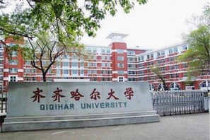 Een bord voor Qiqihar Universiteit, met een rood en wit gebouw op de achtergrond.