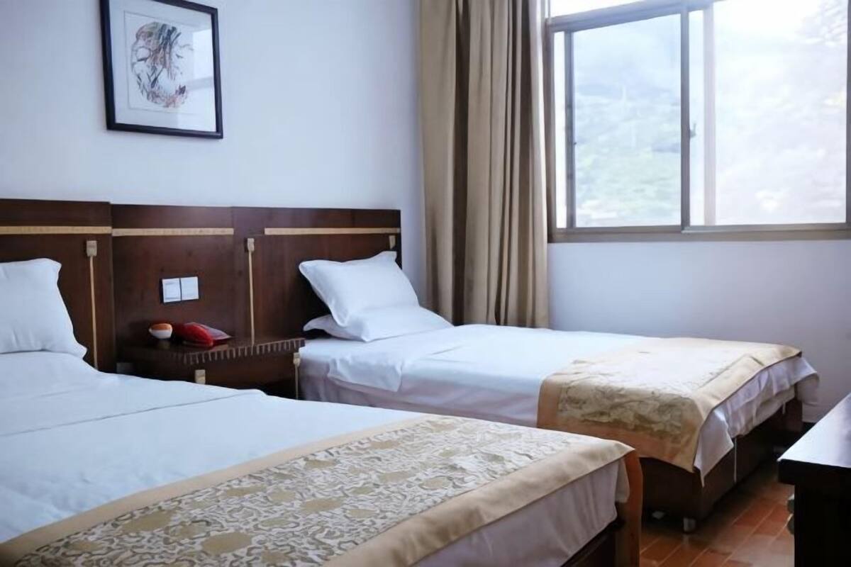A hotel room with two beds, a window with curtains, a framed picture on the wall, and a bedside table with a red object.