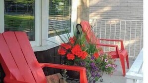 A covered porch with two red Adirondack chairs, a potted plant with red and purple flowers, and a white fence. - Relaxing Birdsong Bungalow (Port Dover)