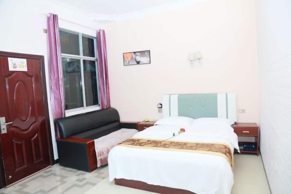 Habitación de hotel con cama, sofá, mesita de noche, ventana con cortinas y un cuadro colgado en la pared.