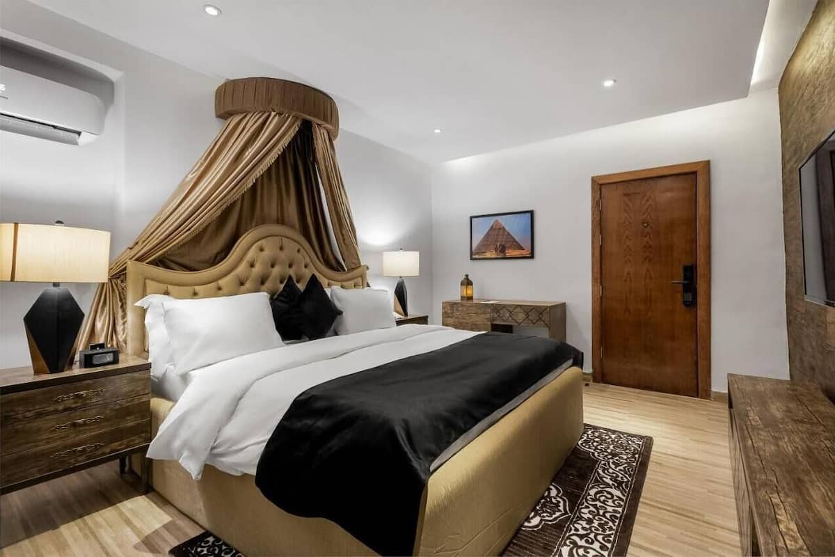 A spacious bedroom with a large bed, a canopy, a wooden nightstand, a bedside table, a lamp, a wooden door, and a framed picture on the wall.