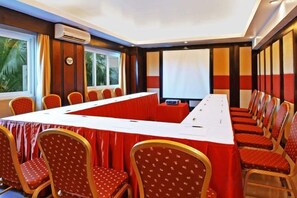 Un salón de conferencias con una larga mesa, sillas, una pantalla de proyector y un ventanal con cortinas.