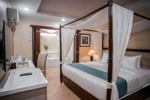 Una cama con dosel y cuatro columnas, una mesita de noche con lámpara, y un escritorio con silla y control remoto.