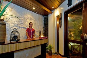 Un área de recepción con un mostrador de madera, una recepcionista, una pared de piedra con esculturas y una puerta de madera grande.