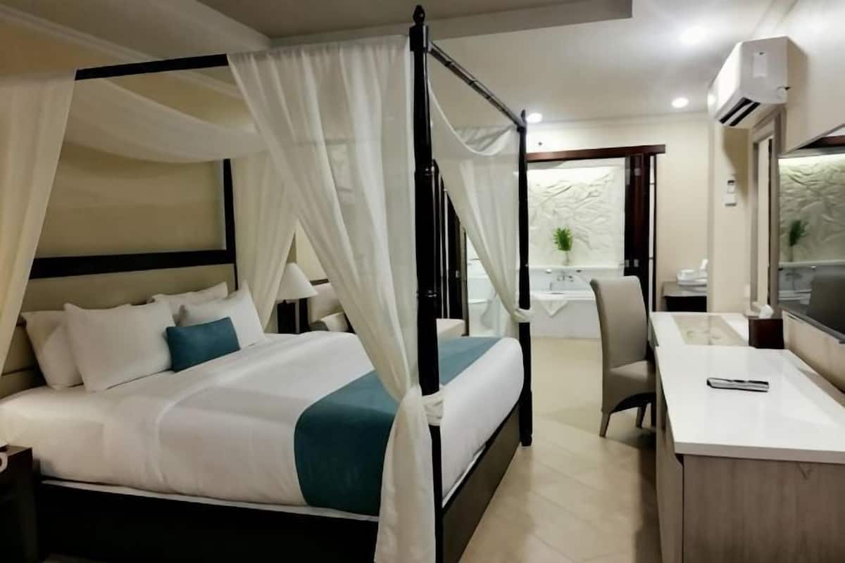 A four-poster bed with white bedding, a desk with a chair, and a small table with a plant.