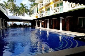 Una piscina con área techada, un edificio con balcones y palmeras al fondo.