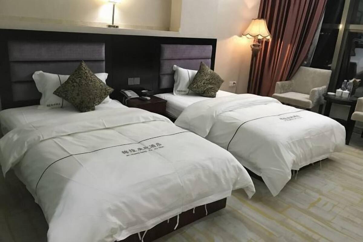 Hotellihuone, jossa on kaksi sänkyä, molemmissa valkoiset vuodevaatteet ja koristeelliset tyynyt, yöpöytä lampun kanssa ja yksi tuoli.
