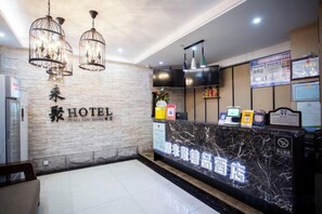 Zona de recepción de un hotel con mostrador de mármol, televisor montado en la pared y techo con luces colgantes.