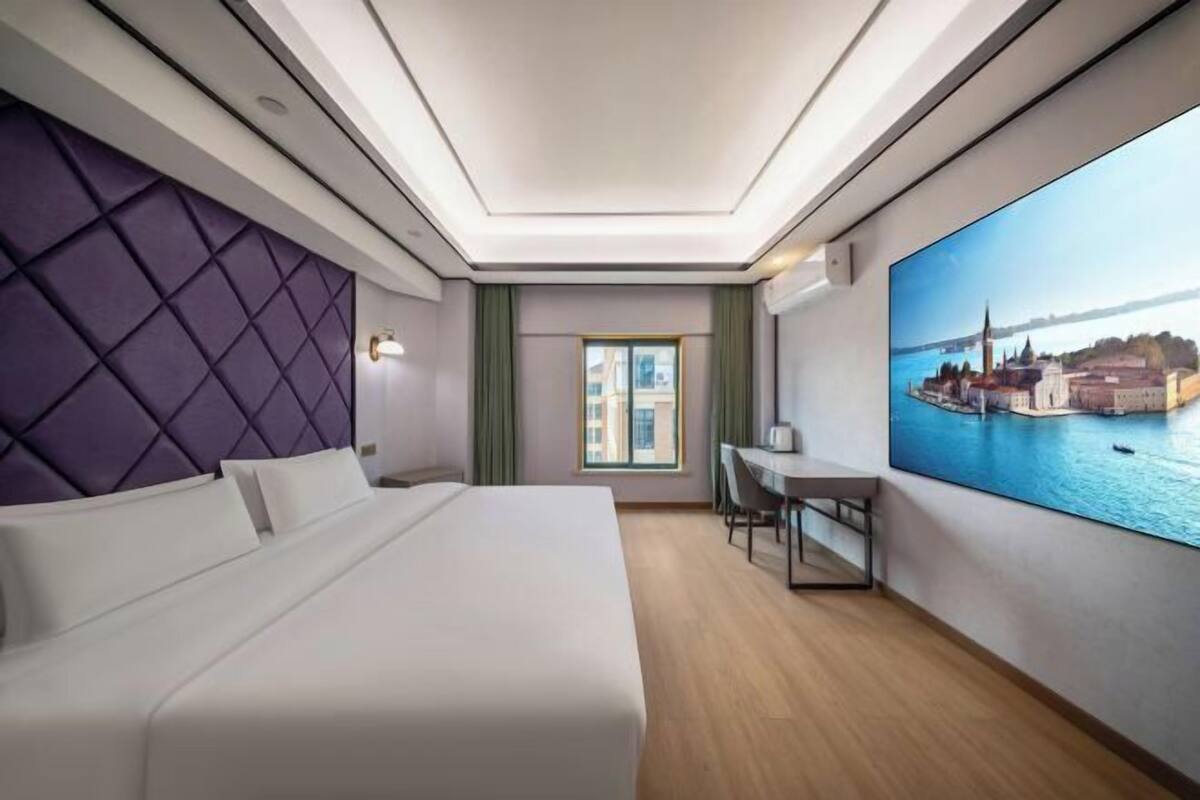 A hotel room with a large bed, a desk, a chair, a window with curtains, and a cityscape painting on the wall.