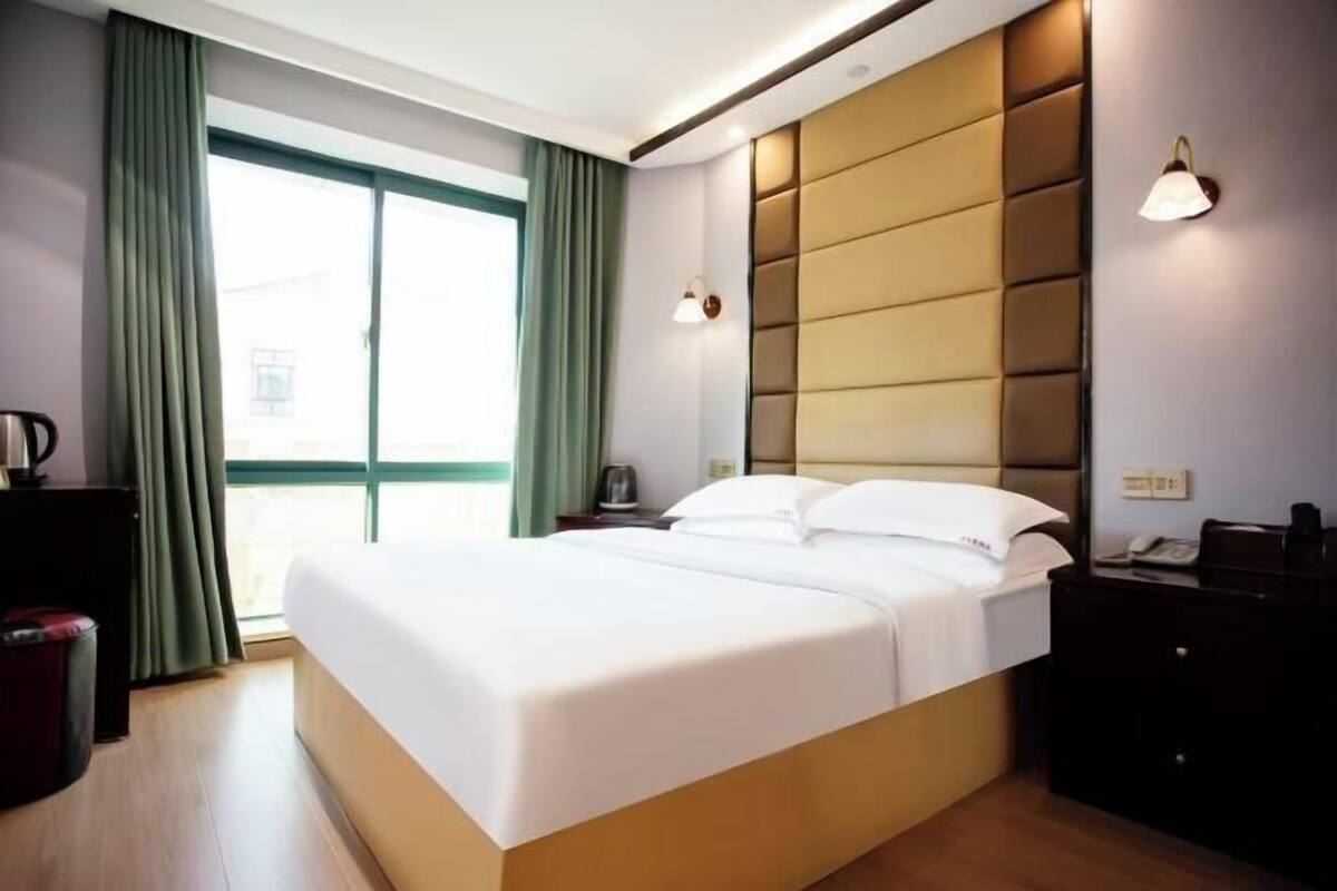 A modern hotel room with a large bed, a bedside table, a nightstand, and a window with curtains.
