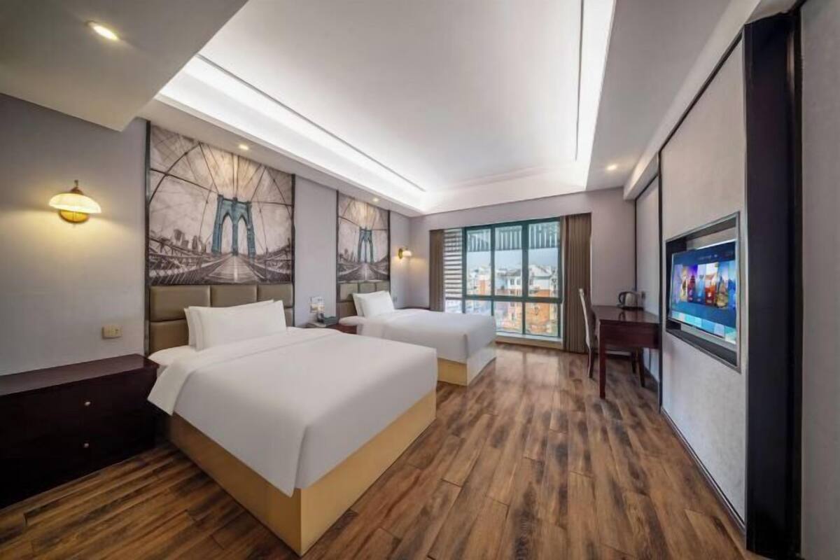 A hotel room with two beds, a large window with a city view, a mounted TV, and a painting on the wall.