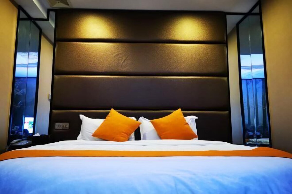 A hotel room with a large bed, a dark headboard, and two windows with curtains.