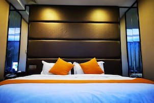 Una habitación de hotel con una cama grande, un cabecero oscuro y dos ventanas con cortinas.