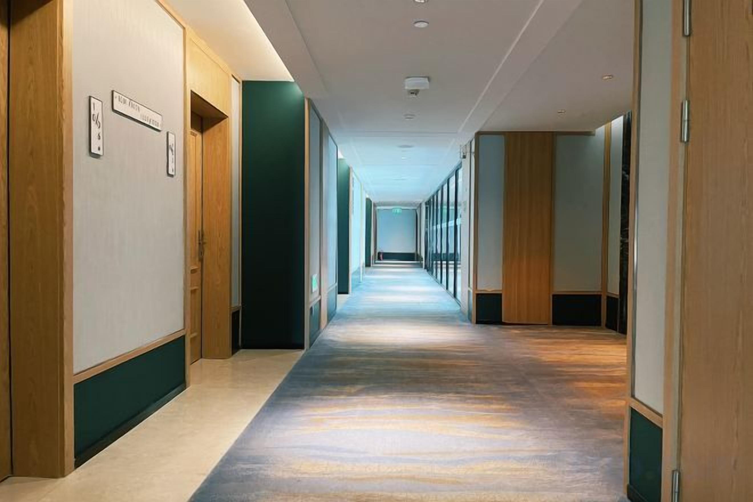 Un corridoio d'albergo con porte su entrambi i lati, pavimento rivestito di moquette e un lungo corridoio che conduce a una stanza luminosa in fondo.
