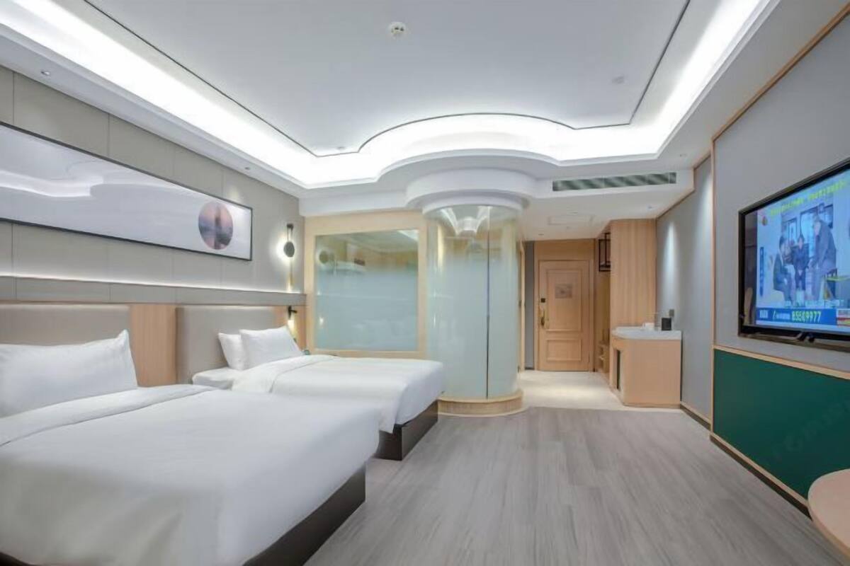 A modern hotel room with two beds, a glass partition, a small bathroom, and a TV screen on the wall.