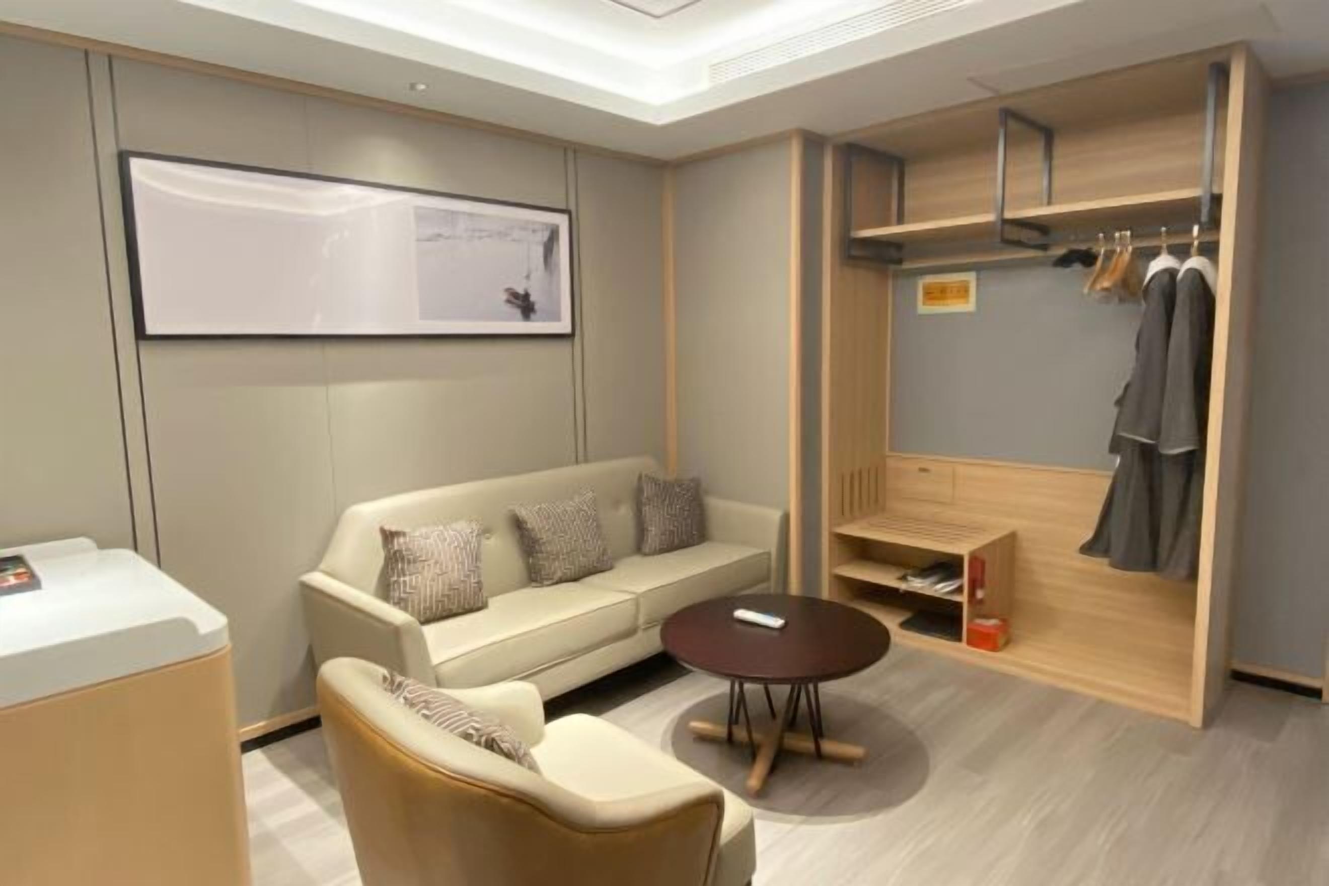 A modern hotel room with a sofa, a coffee table, a wardrobe with hanging clothes, and a framed picture on the wall.