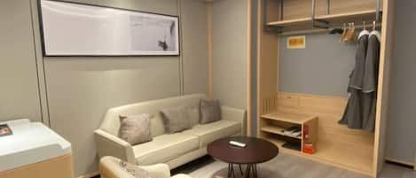 A modern hotel room with a sofa, a coffee table, a wardrobe with hanging clothes, and a framed picture on the wall.