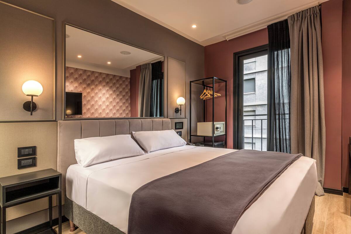 A modern hotel room with a large bed, a bedside table, a mirror, and a wardrobe.