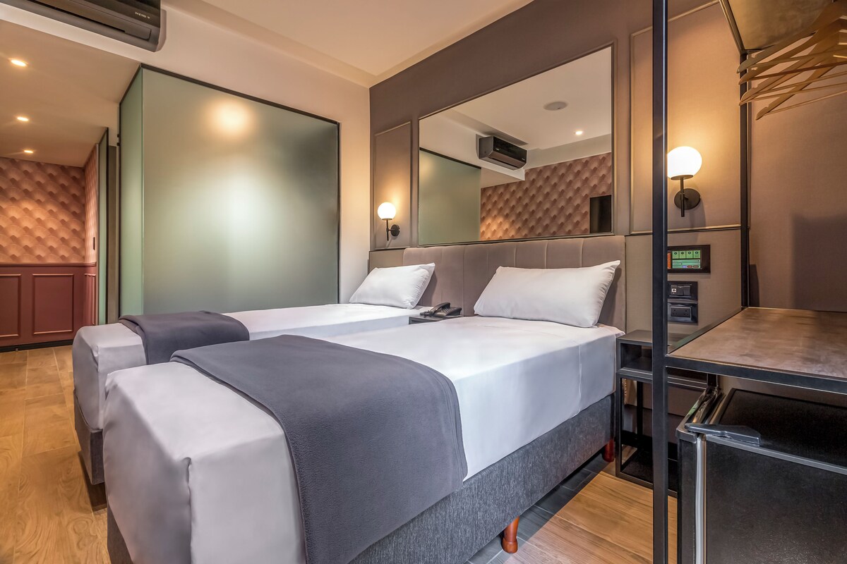 A hotel room with two beds, a desk, a mirror, and a wall-mounted air conditioner.