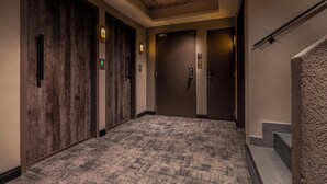 Un couloir avec trois portes, chacune dotée d’un écran numérique et d’un voyant lumineux vert. Le sol est recouvert d’une moquette à motifs et un escalier, avec un mur en pierre sur le côté droit, longe le couloir.