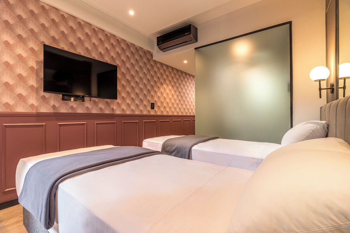 Habitación de hotel con dos camas, un televisor de pantalla plana montado en la pared, un equipo de aire acondicionado y un espejo con luces empotradas.