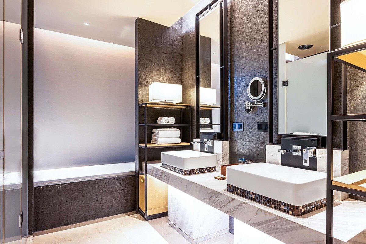 A modern bathroom with two sinks, a large mirror, and a shelf with towels.
