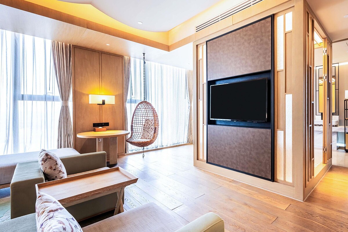 A modern hotel room with a large TV, wooden flooring, a hanging chair, and a dining area.