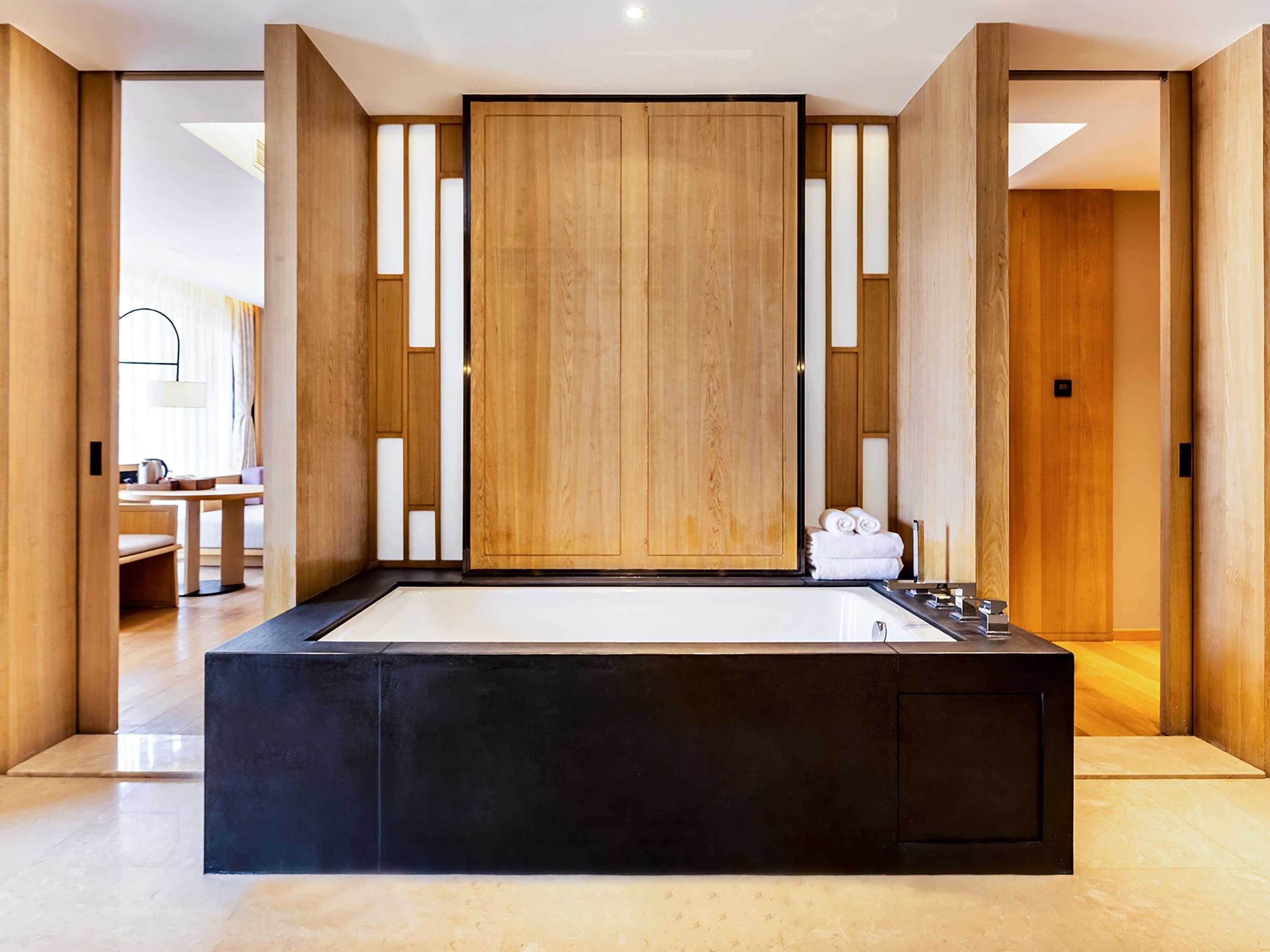 A modern bathroom with a large black bathtub, wooden panels, and a view into a living area.