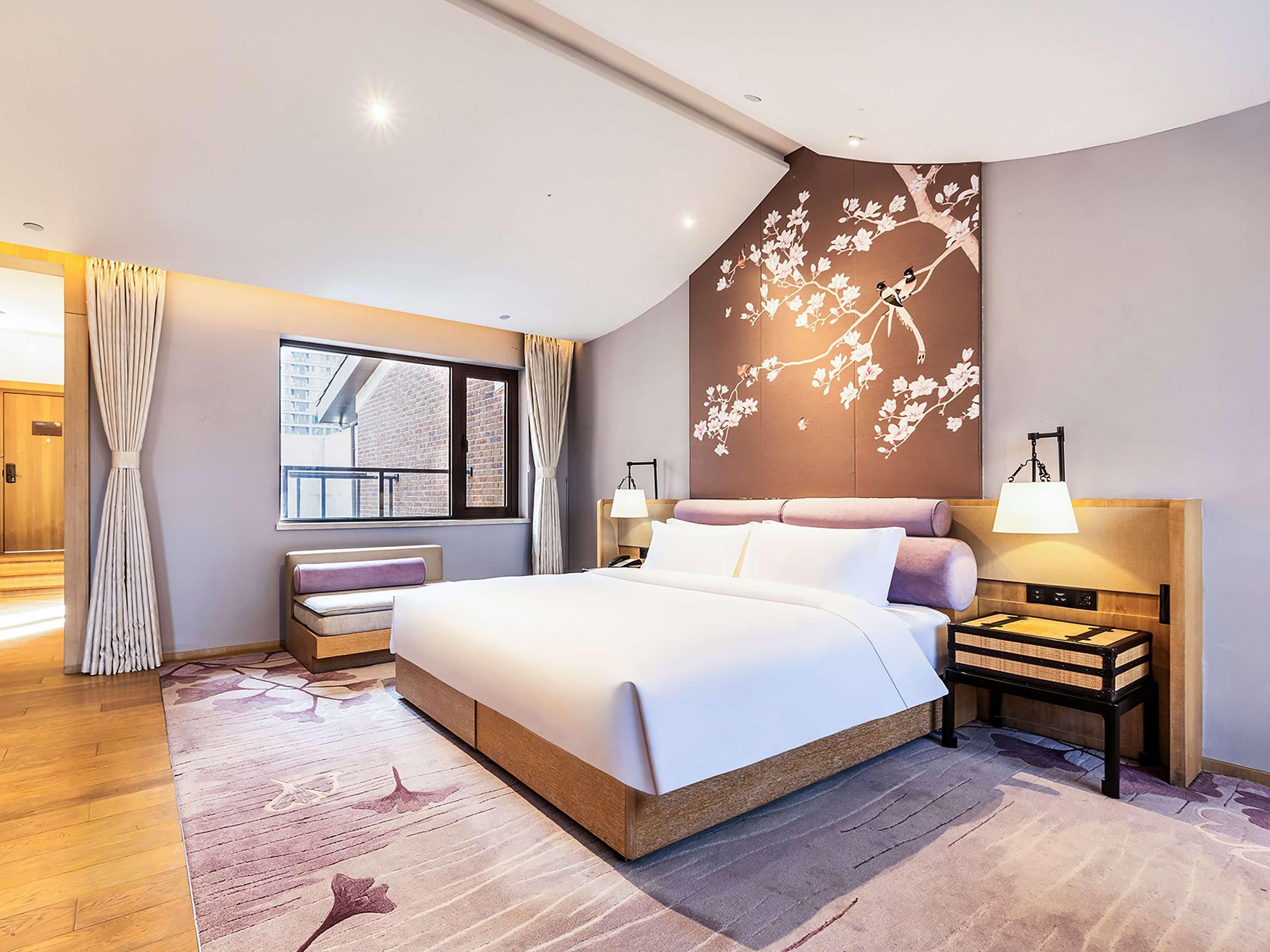 A modern hotel room with a large bed, a bedside table with a lamp, a window with curtains, and a wall mural.