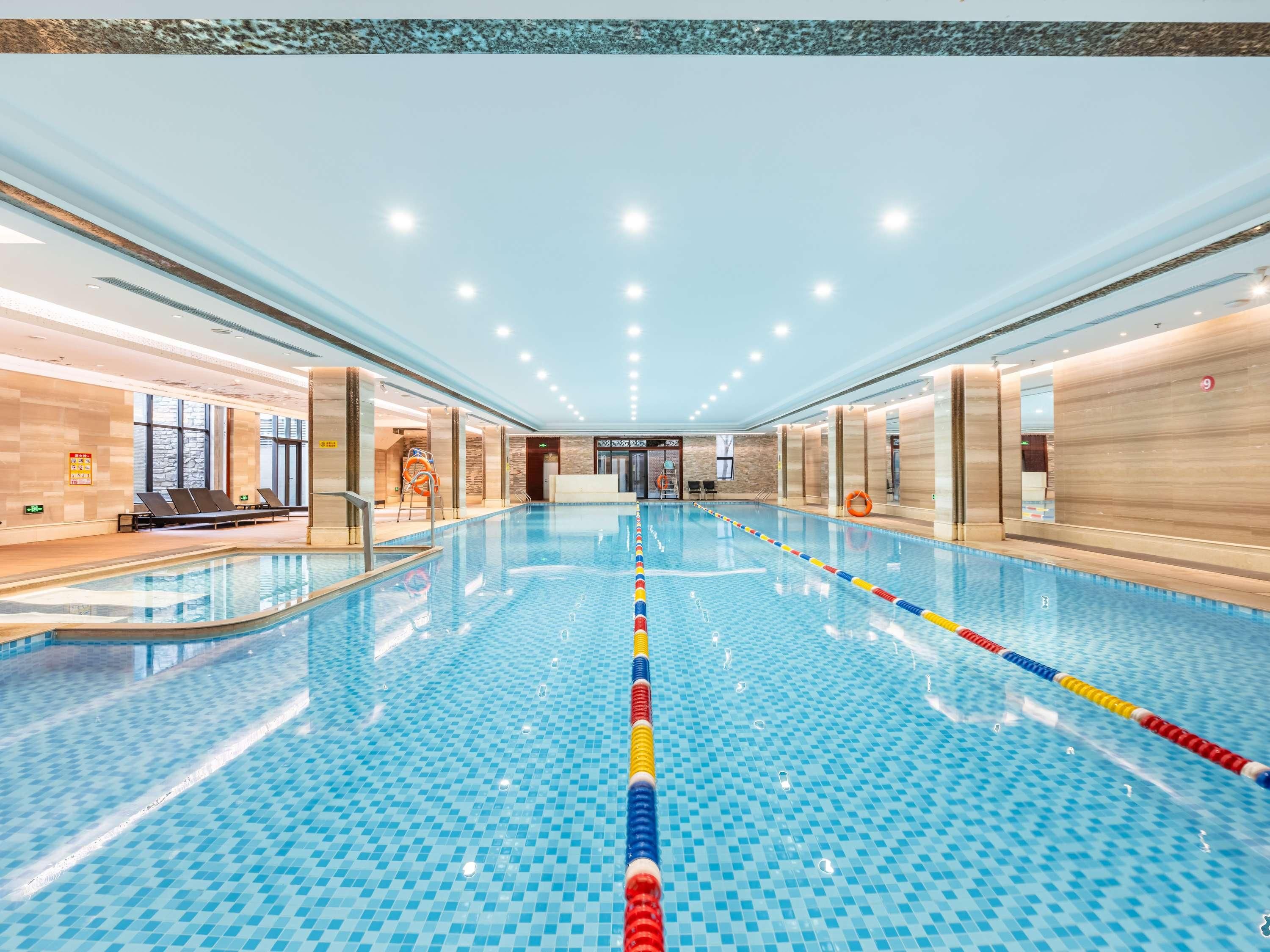 A large indoor swimming pool with clear lane dividers, a lifeguard chair, and a reception area with a sign indicating the pool's location.