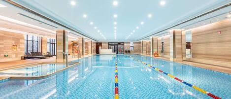 A large indoor swimming pool with clear lane dividers, a lifeguard chair, and a reception area with a sign indicating the pool's location.