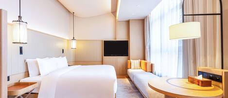 A modern hotel room with a large bed, a bedside table, a small sofa, a round table, and a TV mounted on the wall.